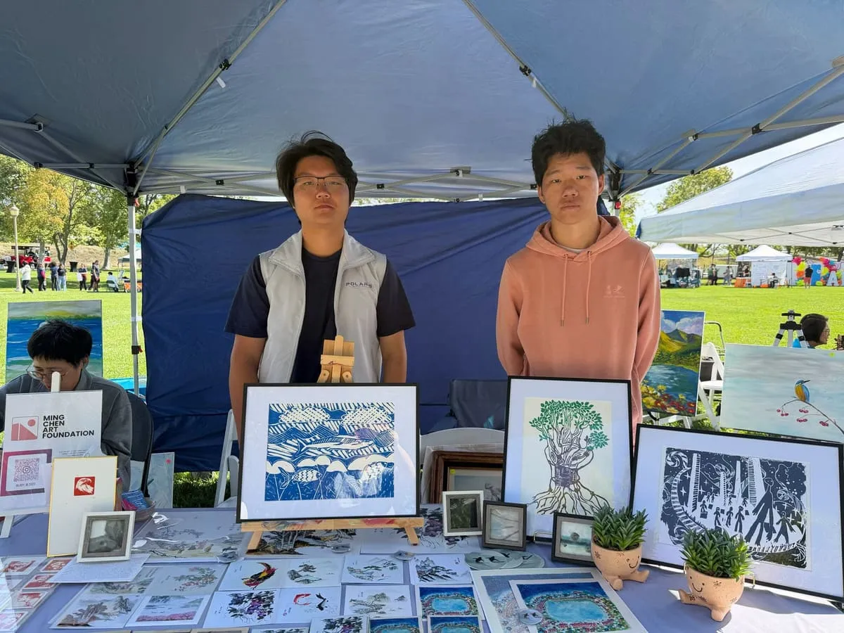 Two teenagers run a sale table of framed prints under a canopy.