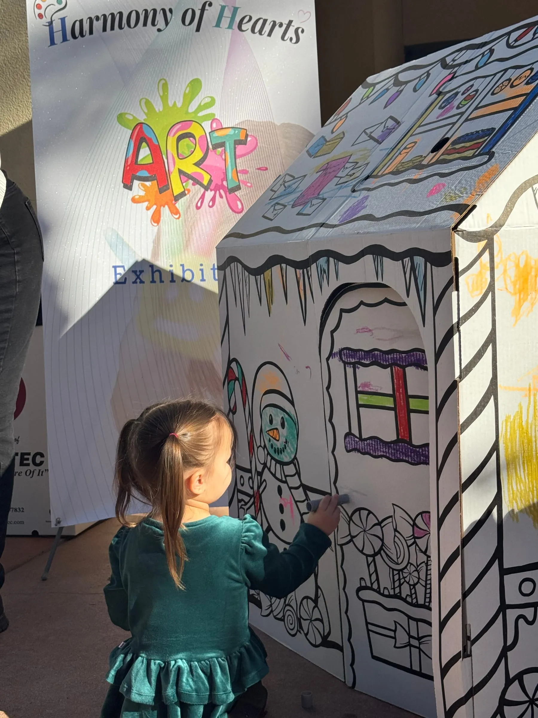 A toddler colors a giant cardboard house beside the Harmony of Hearts banner.